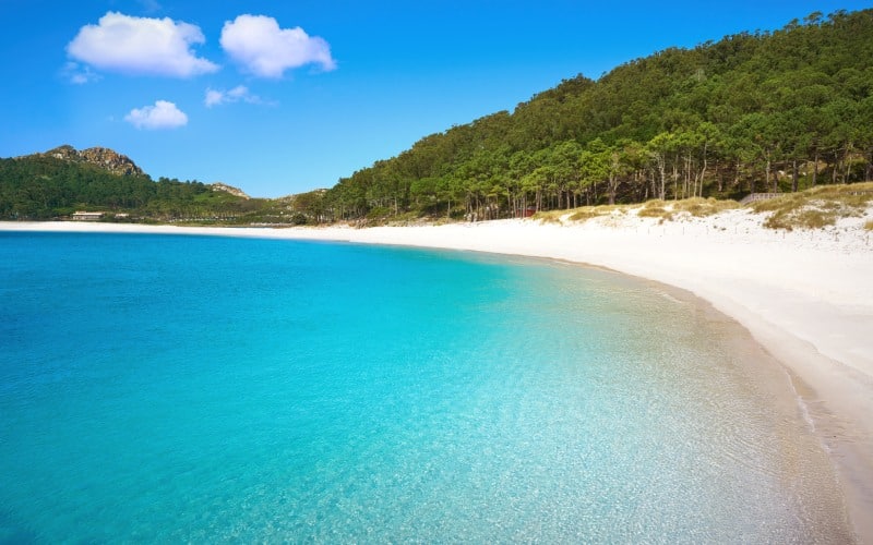 A beach with turquoise waters, and white sand, surrounded by forests