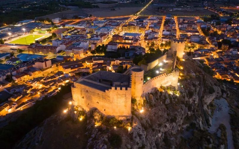 A view from above of a castle by night, lit up on top of a cliff