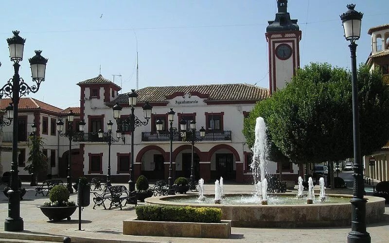 A square with a fountain, a tree and benches, the town hall in the background. 