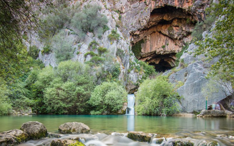 A lake in front of a wall of rock with the entry to a cave and a small waterfall