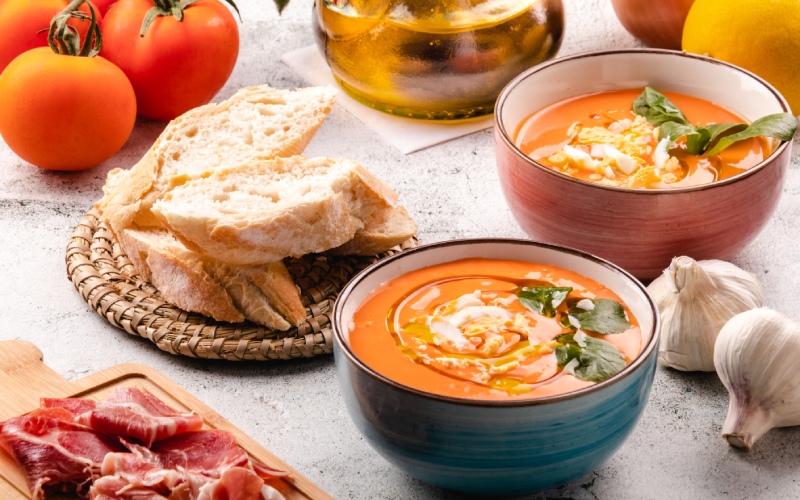 Two coups with orange soups on a table surrounded by other products, like garlic cloves and tomatoes 